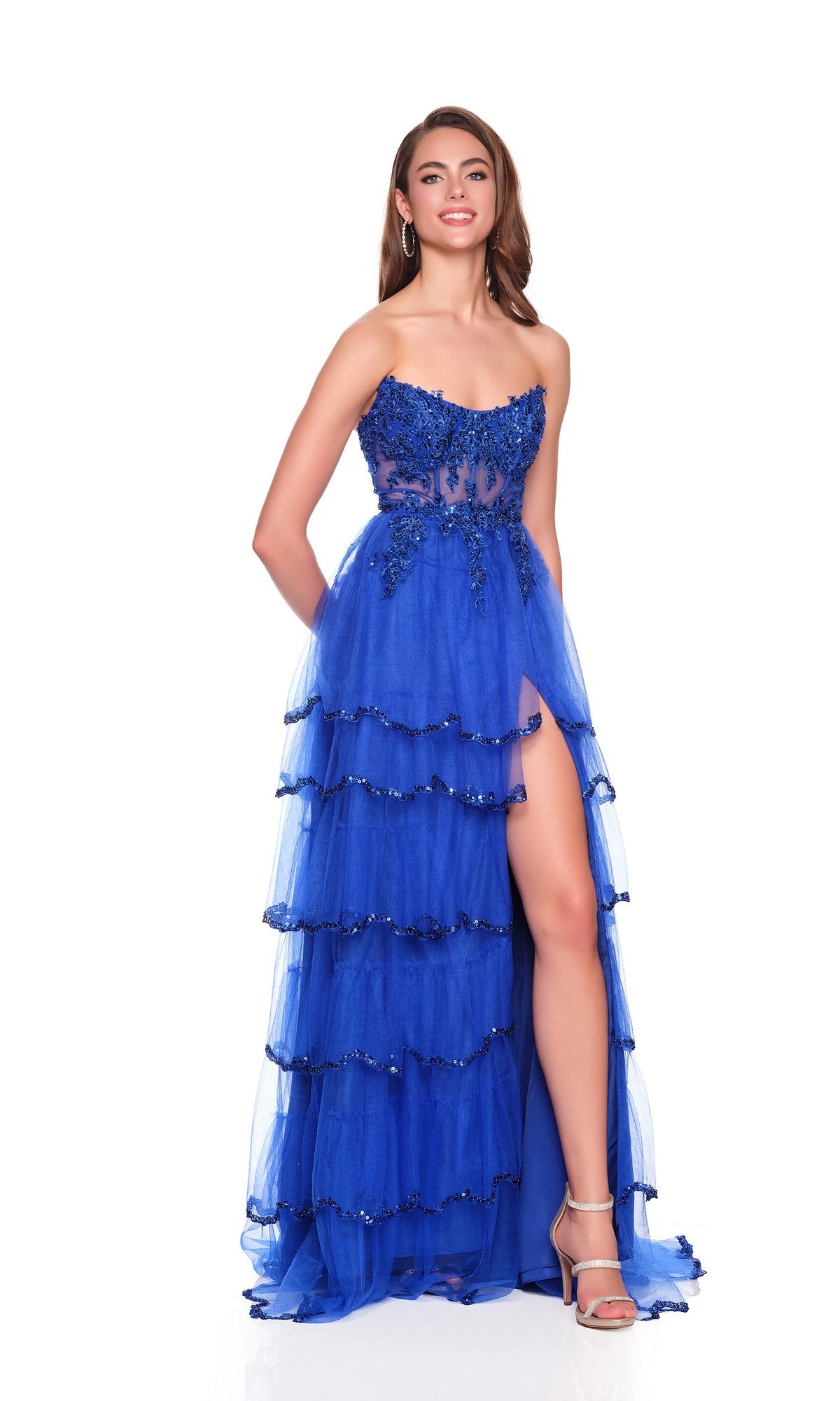 A woman smiles against a white background wearing the Dave and Johnny 12871 long prom dress, a strapless blue gown with a tiered tulle A-line skirt, floral embellishments, and a thigh-high slit.